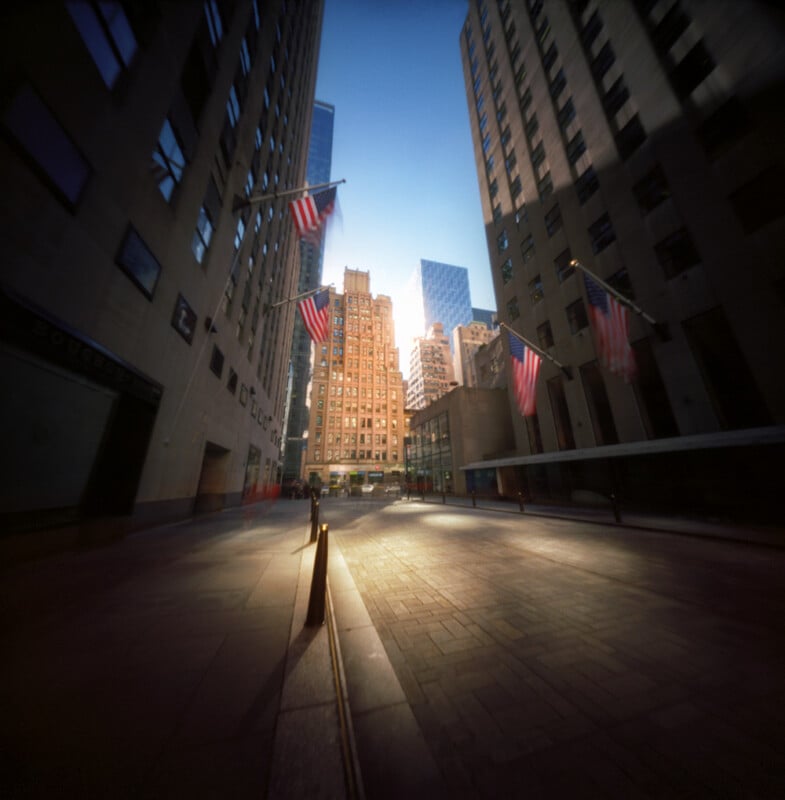 A low-angle view looking down a narrow city street lined with tall buildings and American flags, with sunlight glowing between the structures.