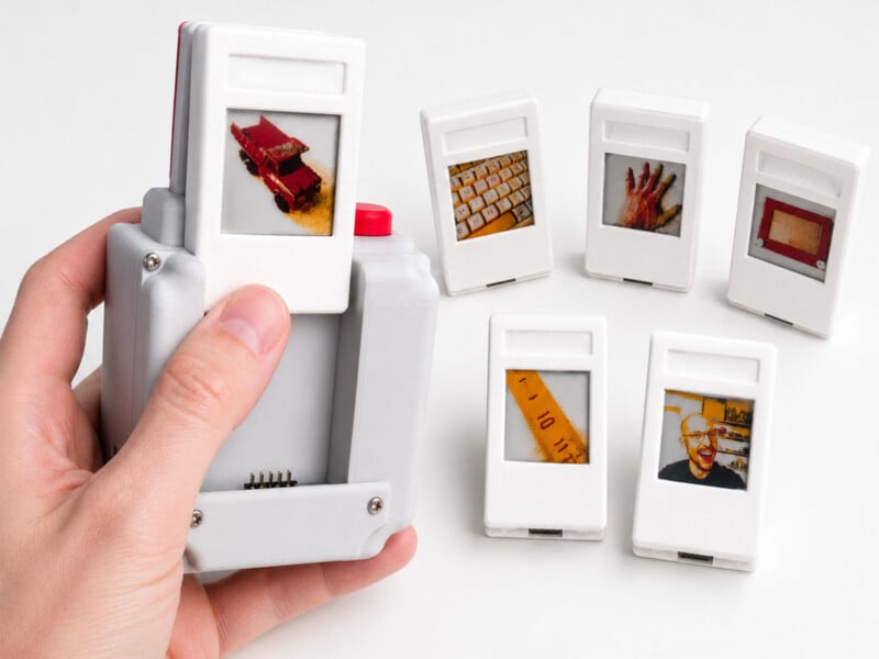 A hand holds a grey, 3D-printed device with a white cartridge inserted, while five additional cartridges featuring various images sit on a white surface.
