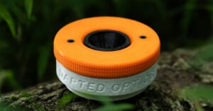 A 3D-printed, two-tone orange and white circular device with the words "ADAPTED OPTICS" embossed on the side, resting on moss.