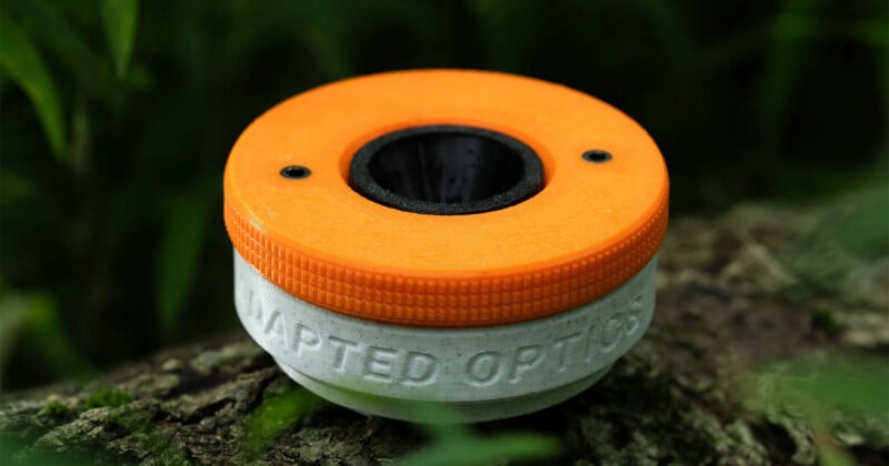 A 3D-printed, two-tone orange and white circular device with the words "ADAPTED OPTICS" embossed on the side, resting on moss.