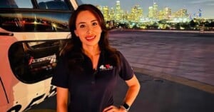 A woman in a dark polo shirt with a "Telemundo 52" logo stands in front of a helicopter with a city skyline at night behind her.