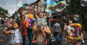 A child is held up by an adult amidst a crowd on a city street filled with many large, colorful floating bubbles.