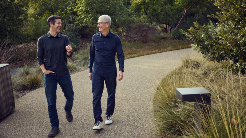 Two men walk along a gravel path in a park, smiling and looking at each other while wearing button-down shirts and jeans.
