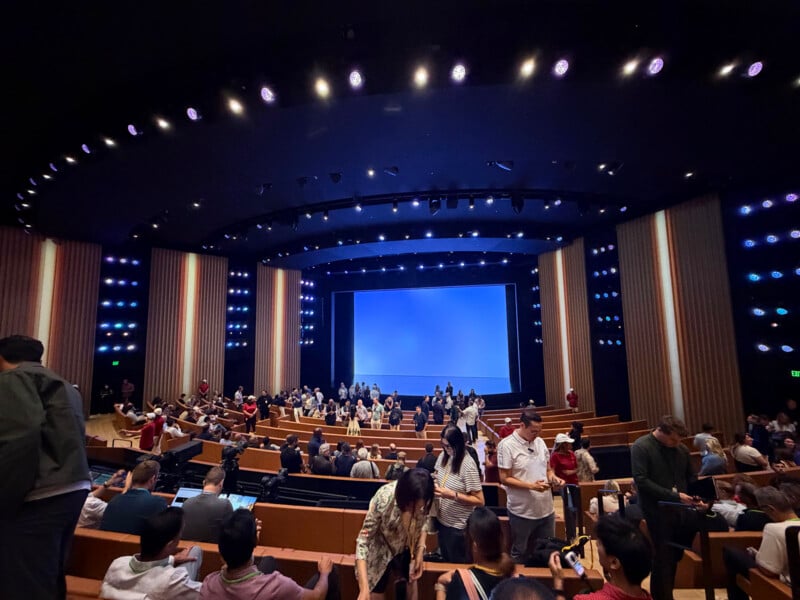 A large, modern auditorium with tiered seating is filled with people facing a large, blank blue projection screen on stage.