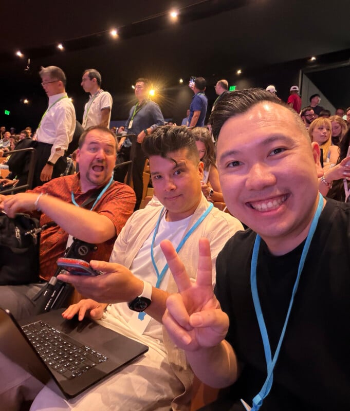 Three men sit in an auditorium, with the man on the right smiling and making a peace sign while the others look toward the camera.