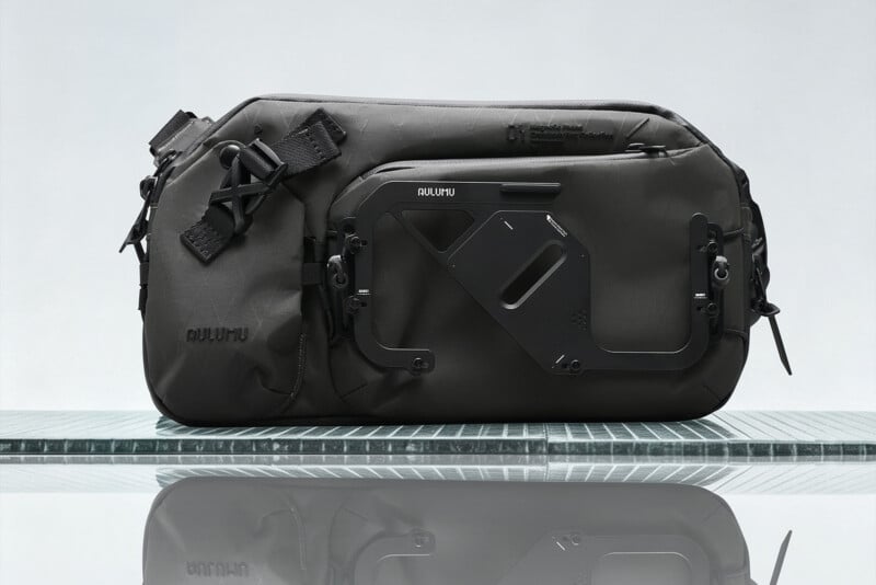 A dark gray AULUMU crossbody bag with a prominent black metal magnetic phone mount attached to the front, resting on a reflective surface.