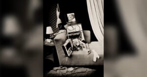 A disassembled computer tower sits on a sofa, with a laptop displaying a woman's face resting on top, near a bra hanging on the wall.