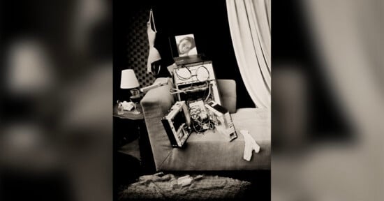 A disassembled computer tower sits on a sofa, with a laptop displaying a woman's face resting on top, near a bra hanging on the wall.