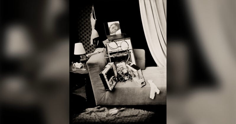 A disassembled computer tower sits on a sofa, with a laptop displaying a woman's face resting on top, near a bra hanging on the wall.