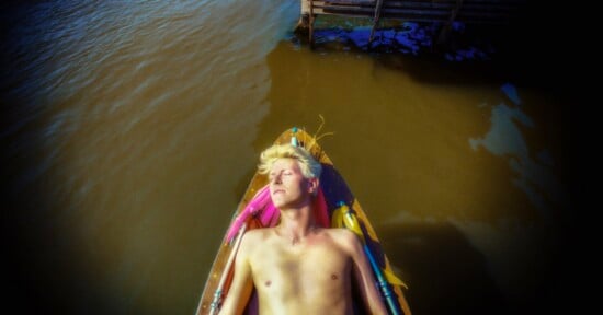A shirtless person with blonde hair lies with their eyes closed in a small wooden boat floating on murky water.