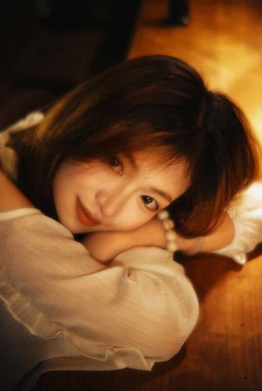 A young woman with shoulder-length brown hair rests her head on her folded arms, looking directly at the camera in warm, soft lighting.