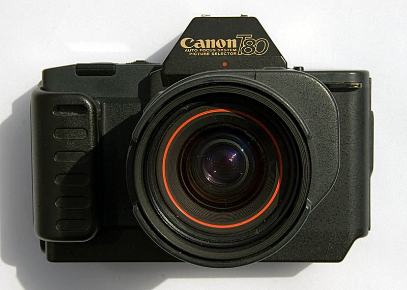 A black Canon T80 35mm film camera with an attached lens, viewed from the front against a white background.