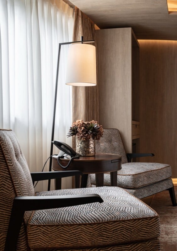 Two patterned armchairs face a small round wooden table holding a telephone and a vase of flowers, illuminated by a floor lamp.