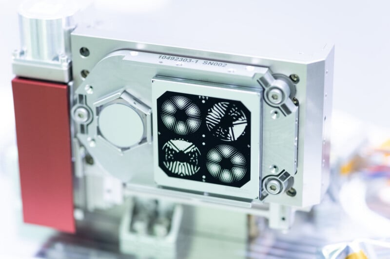 A precision mechanical component featuring a square plate with four circular, patterned optical elements, labeled "10492303-1 SN002.