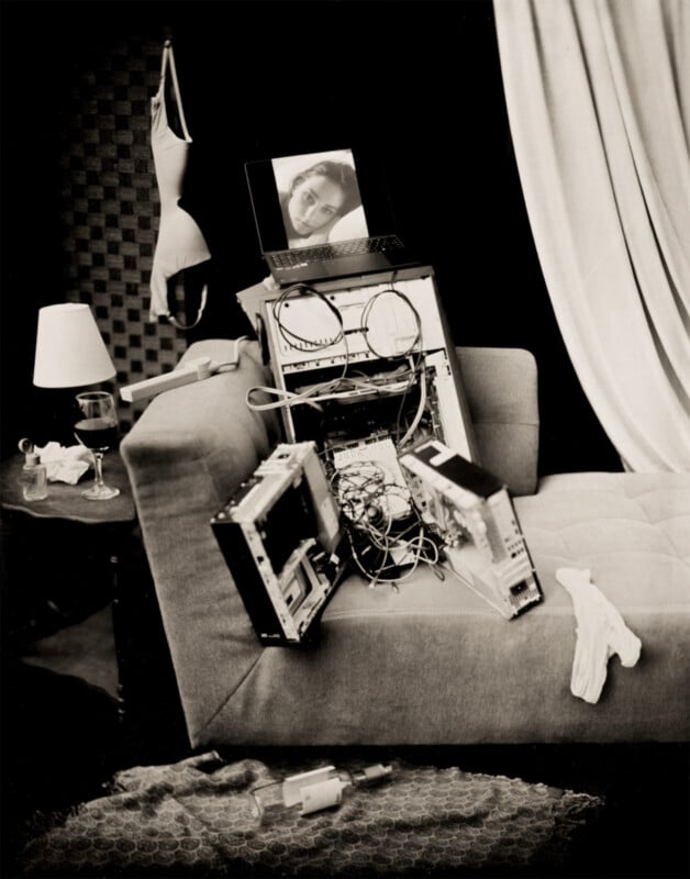 A disassembled computer tower sits on a sofa, topped with a laptop displaying a woman's face, near a side table with a lamp, wine glass, and hanging lingerie.