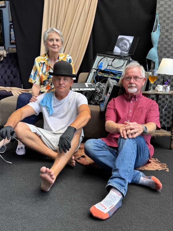 Three people sit in a room with a disassembled computer tower, a laptop displaying a portrait, and a lamp on a side table.