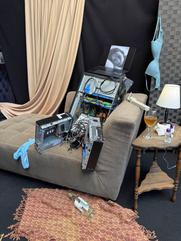 A disassembled computer tower sits on a brown chaise lounge, topped with a laptop displaying a black-and-white portrait, with a bra hanging nearby and a wine glass on a side table.