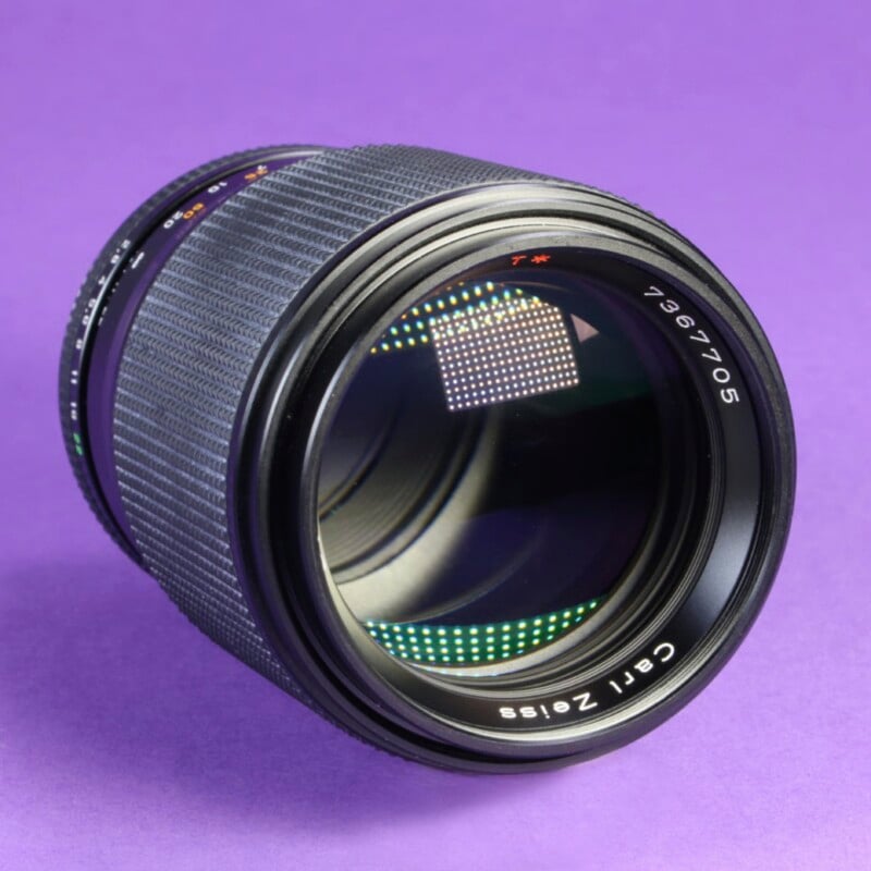 A black Carl Zeiss camera lens with a serial number 7367705 sits on a solid purple background, showing a reflection of a grid pattern on its front element.