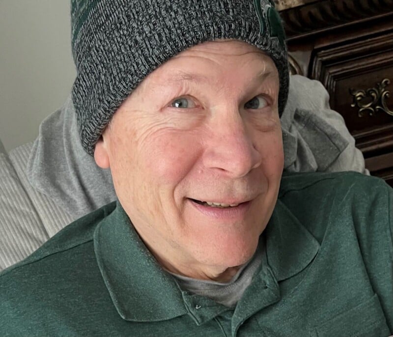 A smiling man with light skin and blue eyes wears a gray and green knit beanie and a green polo shirt.