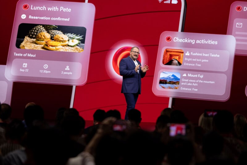 A man in a blue suit stands on a stage in front of a large red screen displaying floating digital interface panels with text and images.