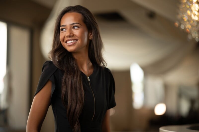 A smiling woman with long, dark hair wears a black dress with a front zipper, looking off to the side in a softly lit room.