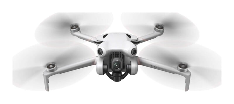 A white DJI Mini 4 Pro drone is shown from the front with its propellers spinning, featuring a central camera gimbal and forward-facing sensors.