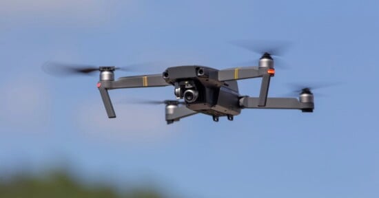 A gray quadcopter drone with spinning propellers flies against a clear blue sky.