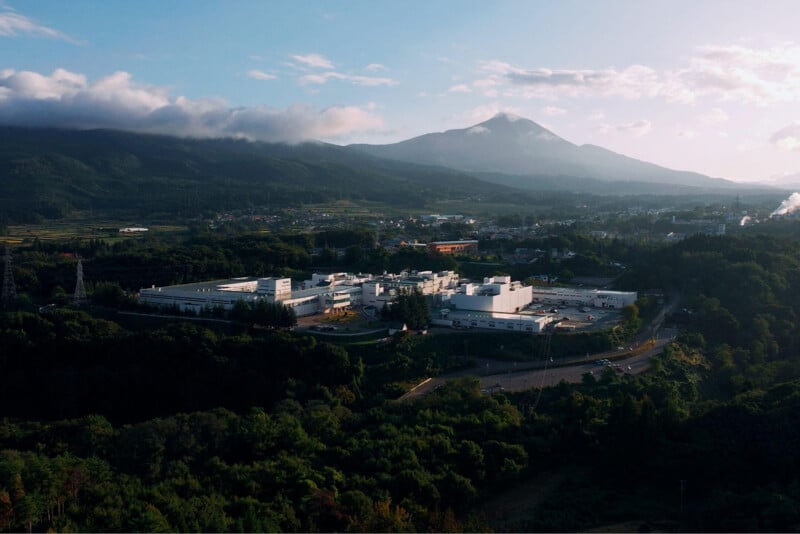 An aerial view shows a large white industrial complex nestled in a forested landscape beneath a prominent mountain peak.