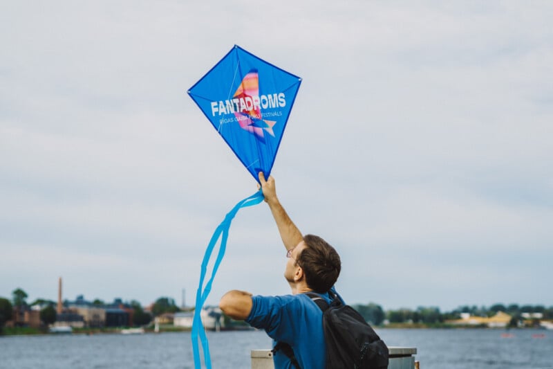 A person with a backpack stands by a body of water, holding a blue diamond-shaped kite with the text "FANTADROMS" printed on it.