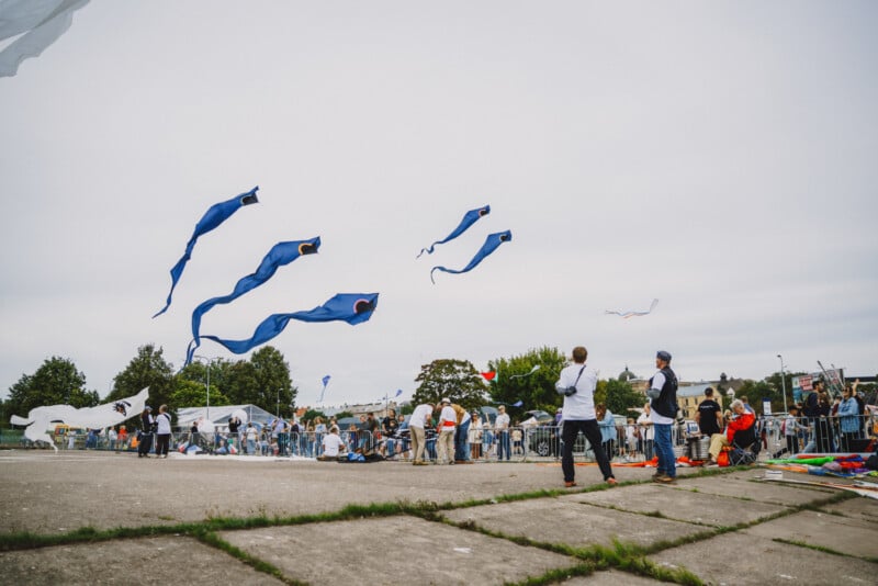 People gather on a paved lot to fly several long, blue, fish-shaped kites against a cloudy sky.