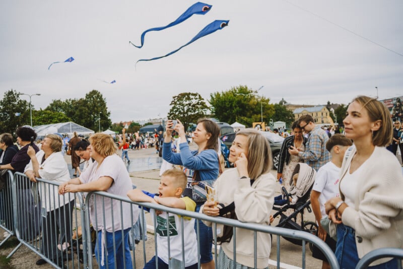 A crowd of people stands behind a metal barrier, looking up at several blue kites flying in the sky.