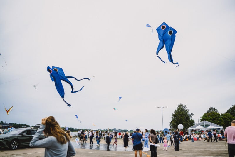 People gather in a parking lot to fly large, blue, octopus-shaped kites under an overcast sky.