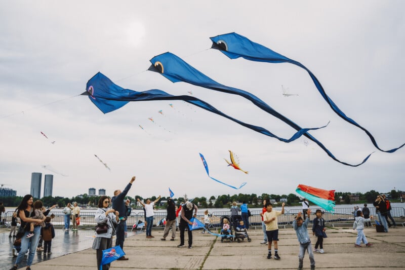 A group of people fly various kites, including three long blue tube kites, on a paved waterfront area under a cloudy sky.