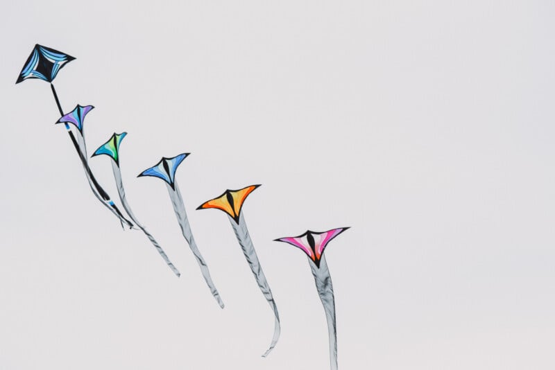 A diagonal line of five colorful, triangular kites with long, flowing tails flies against a plain, light gray sky.