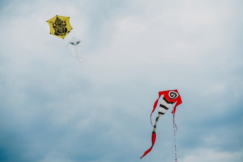 Three kites fly against a cloudy sky: a yellow kite with a Ganesha design, a white kite with eye graphics, and a red kite.