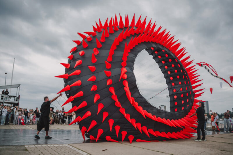 Two people hold the tether lines of a large, circular, black kite covered in red spikes at an outdoor festival.