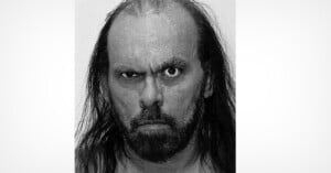 A black-and-white close-up portrait of a man with long dark hair, a beard, and an intense, glaring expression.