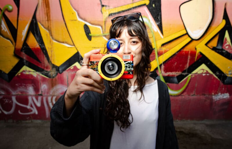 A woman with long dark hair holds a red and yellow fisheye camera in front of her face against a graffiti-covered wall.