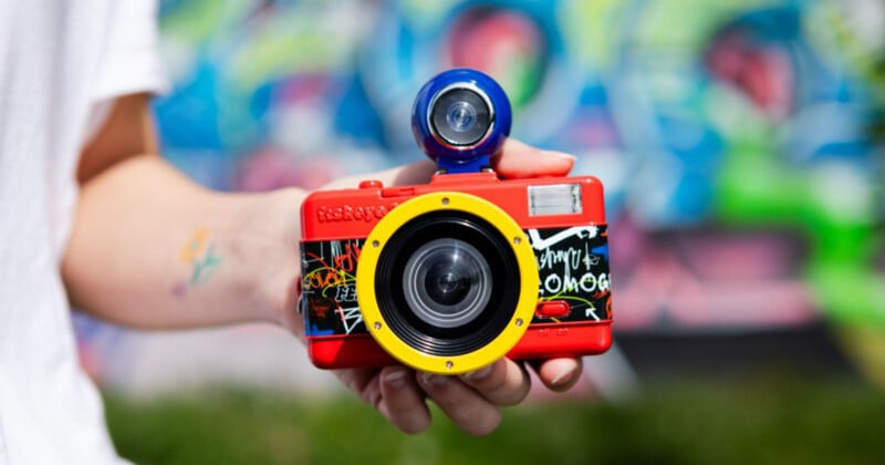 A hand holds a bright red, yellow, and blue fisheye film camera with graffiti-style patterns against a blurred, colorful background.
