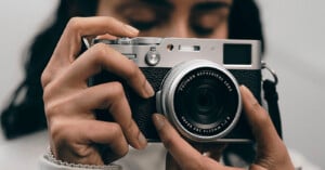 A person holds a silver and black digital camera with a Fujinon 23mm lens up to their face.