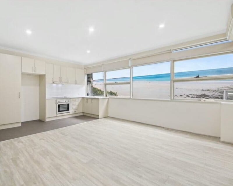 An open-plan kitchen and living area with light wood-look flooring, cream cabinetry, and large windows overlooking a beach and ocean.