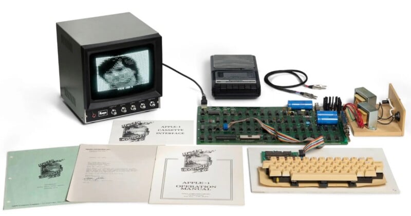 An Apple-1 computer setup including a circuit board, keyboard, monitor displaying a portrait, cassette player, and various manuals on a white background.