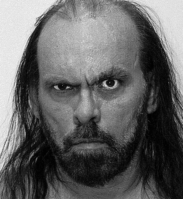 A black-and-white close-up portrait of a man with long, thinning hair and a full beard, looking directly at the camera.