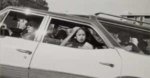 A black-and-white side view of a station wagon with a woman driving, a woman in the back seat adjusting her hair, and two children in the rear cargo area.