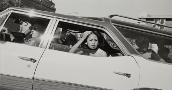 A black-and-white side view of a station wagon with a woman driving, a woman in the back seat adjusting her hair, and two children in the rear cargo area.