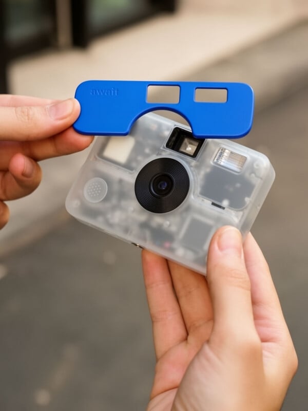 A person holds a translucent digital camera and a separate blue plastic faceplate labeled "await" above it.