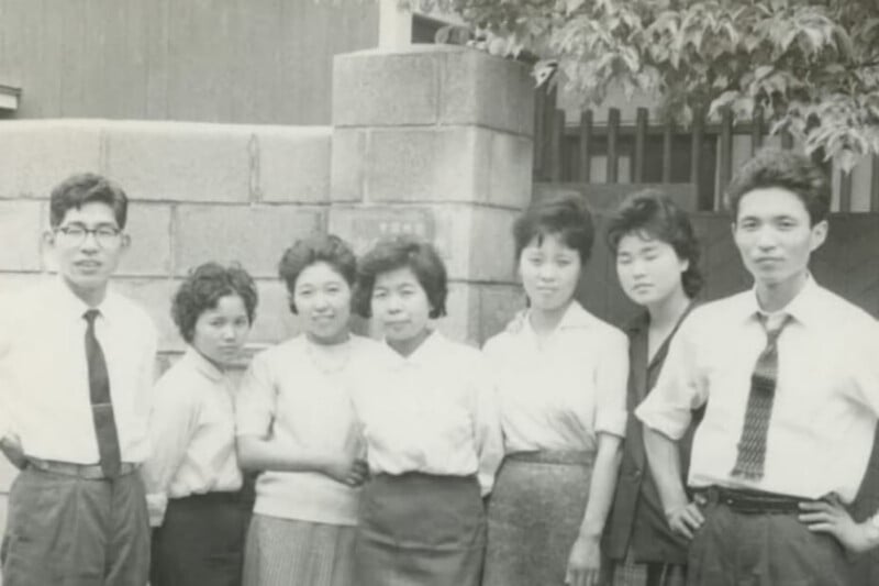A group of seven young adults, two men and five women, stand in a row outdoors in front of a concrete block wall.