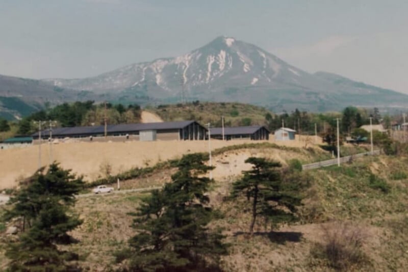 A large mountain with snow-capped peaks rises behind a complex of long, low buildings situated on a cleared hillside.