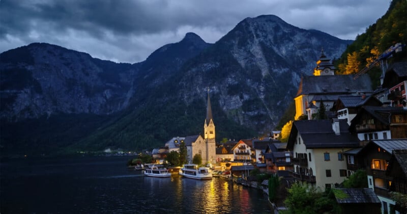 A lakeside village with a tall church spire and illuminated buildings sits at the base of steep, dark mountains at dusk.