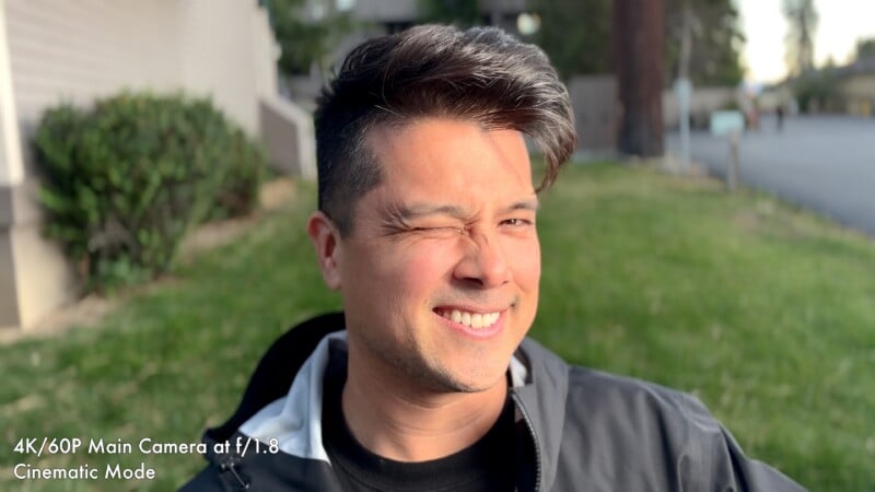 A man with a stylish haircut winks at the camera while standing outdoors in front of a grassy lawn.